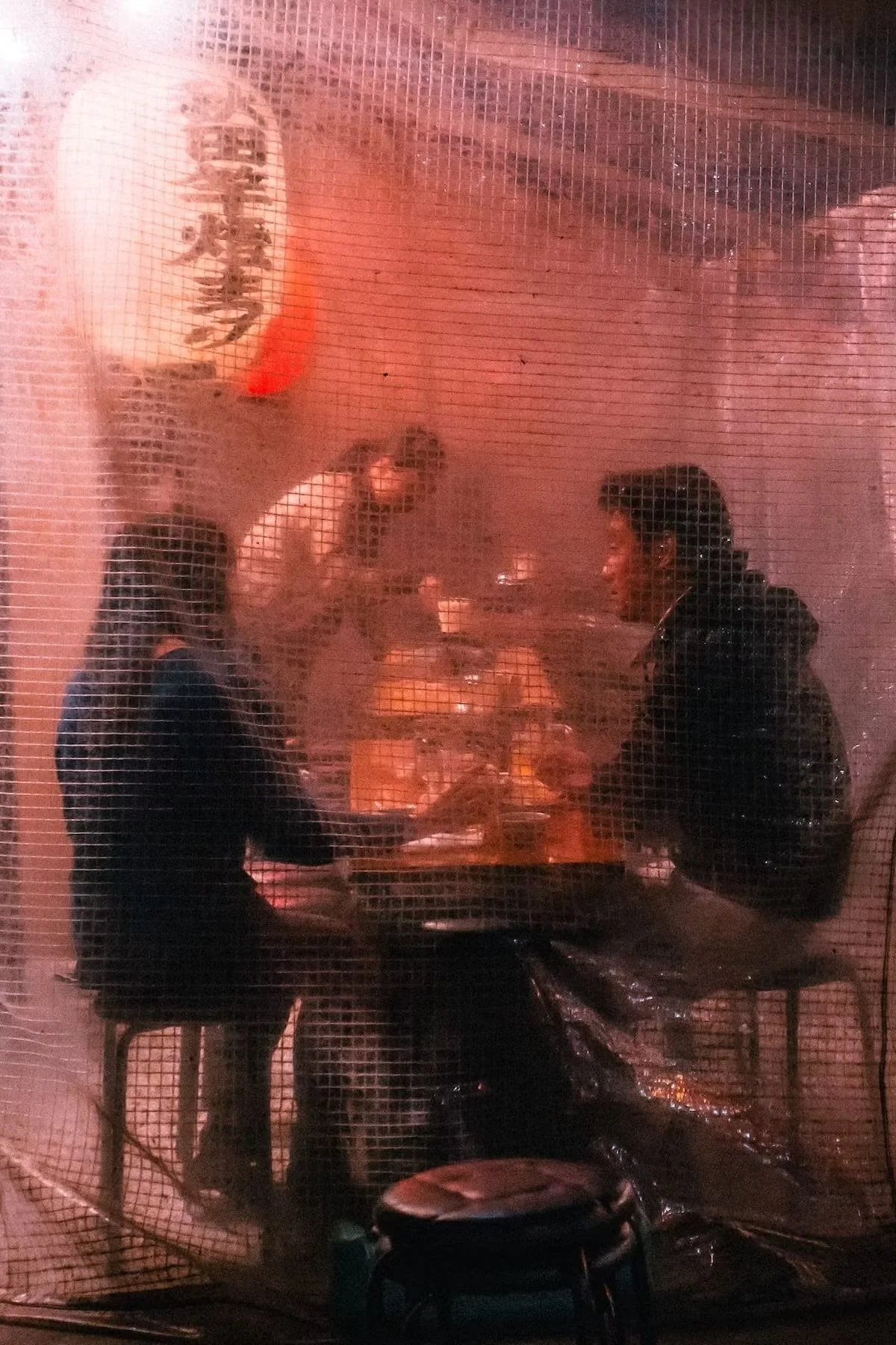 People sitting at a table inside a restaurant, viewed through a window with a mesh screen in an Izakaya in Tokyo.