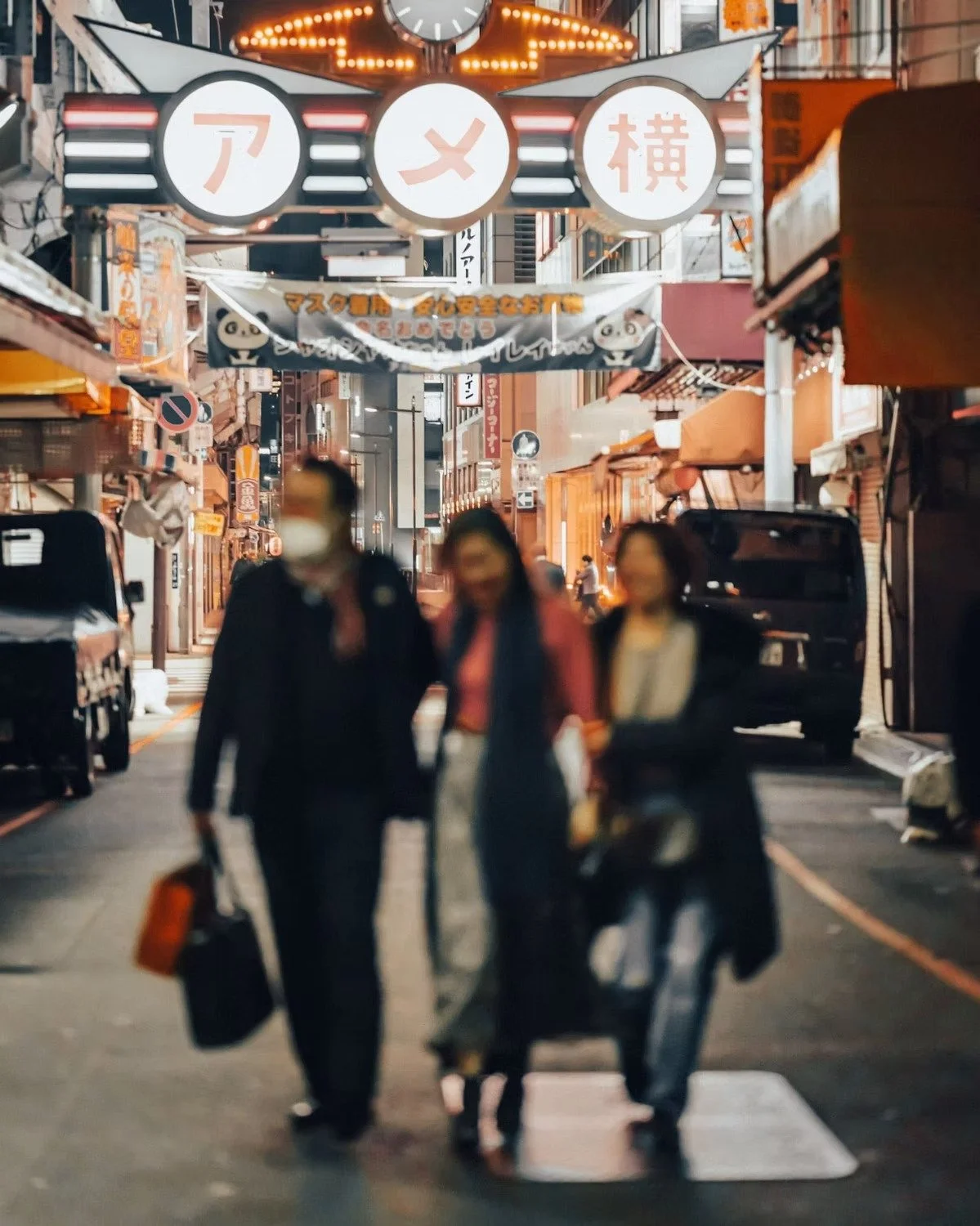 Night scene of a busy street in Japan with illuminated signs and storefronts. Three blurred pedestrians, two women and one man, walk toward the camera in the foreground, with some carrying bags. Cars are parked along the sides of the street, and vari