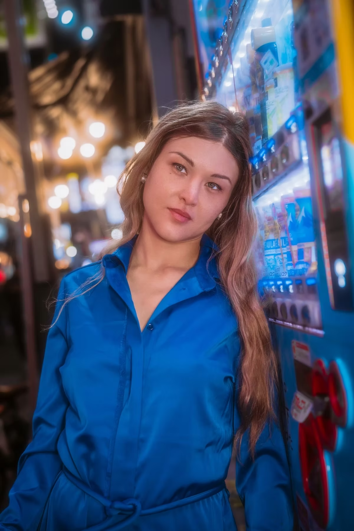 A woman in a blue jumpsuit stands in front of a vending machine in a neon-lit alley in Tokyo, looking confidently at the camera.