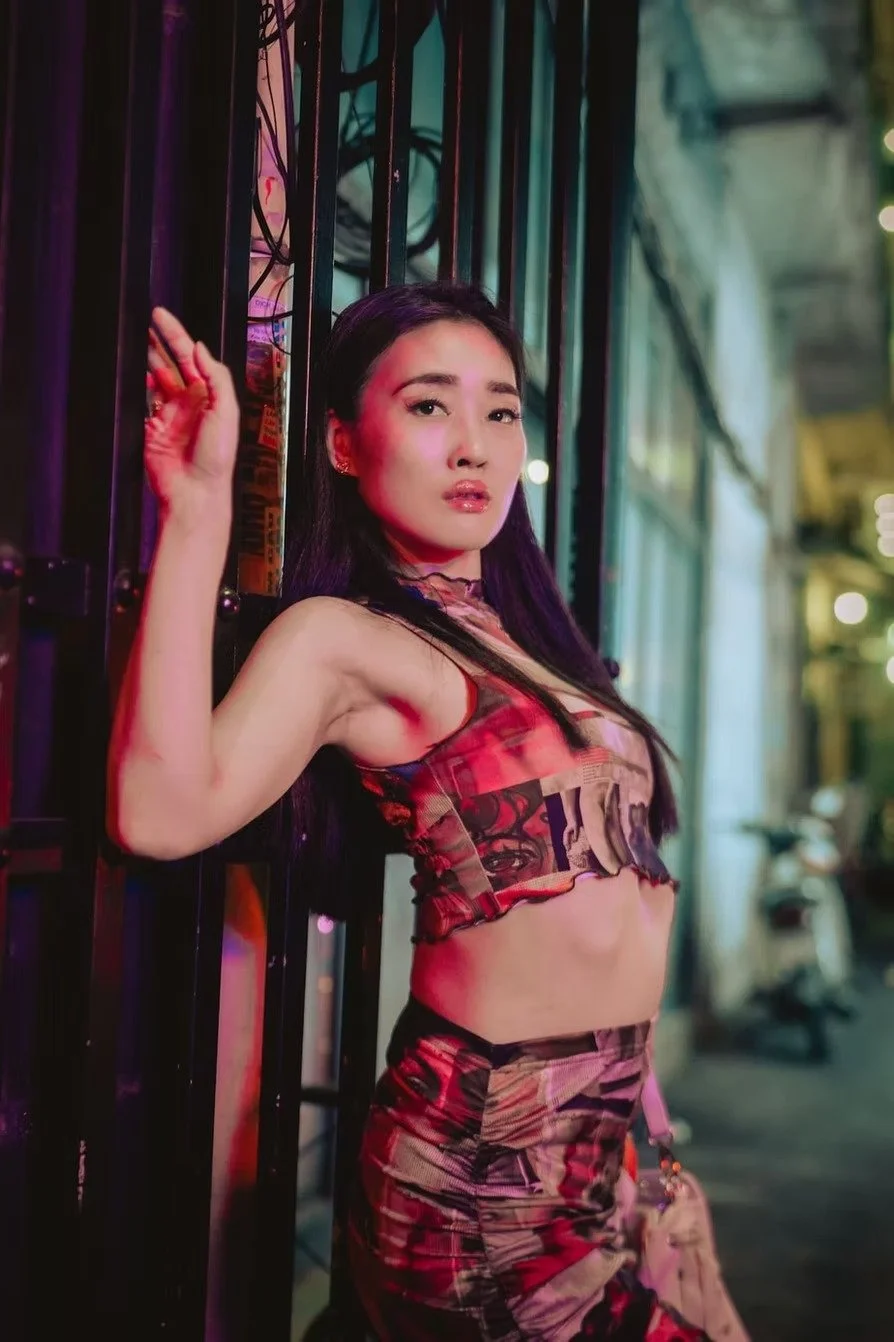 A woman with long dark hair posing against a metal gate with purple and pink neon lighting in the background on the streets of Saigon.