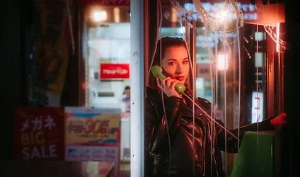 A woman talking on a corded phone in a phone booth at night with neon lights reflecting on the glass
