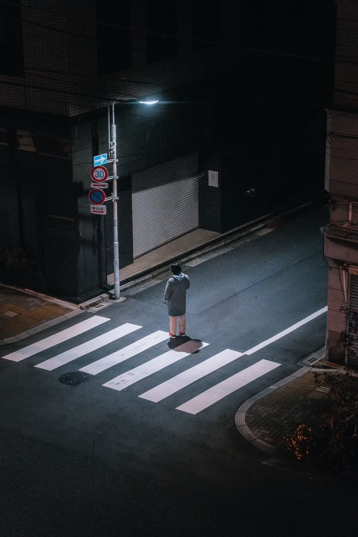 A person standing at a crosswalk at night, illuminated by a streetlight, near a building with a closed metal shutter and street signs in Tokyo.