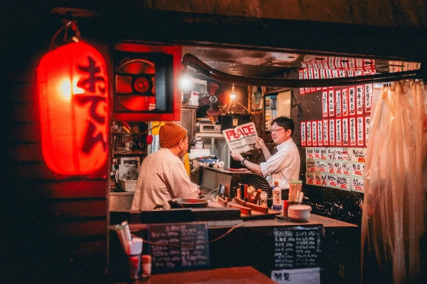 A Japanese restaurant with a red lantern, two men sitting at the counter, and menu signs with Japanese characters on the wall. One man is reading a newspaper in the Nakano Broadway area.