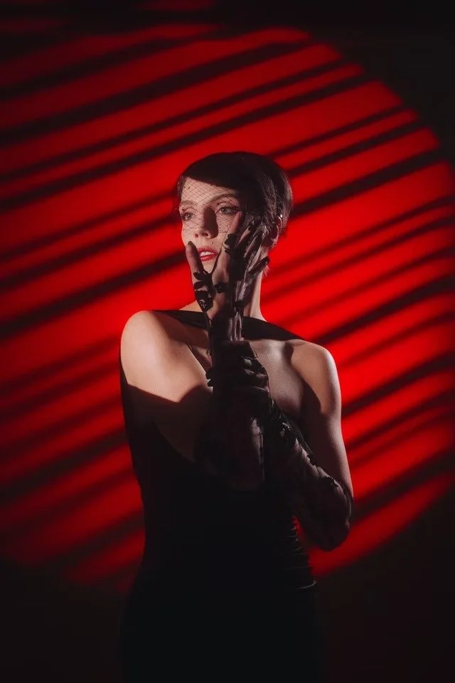 A woman with short dark hair wearing black lace gloves and a black dress, standing against a red background with black striped shadows, holding her hand to her face.