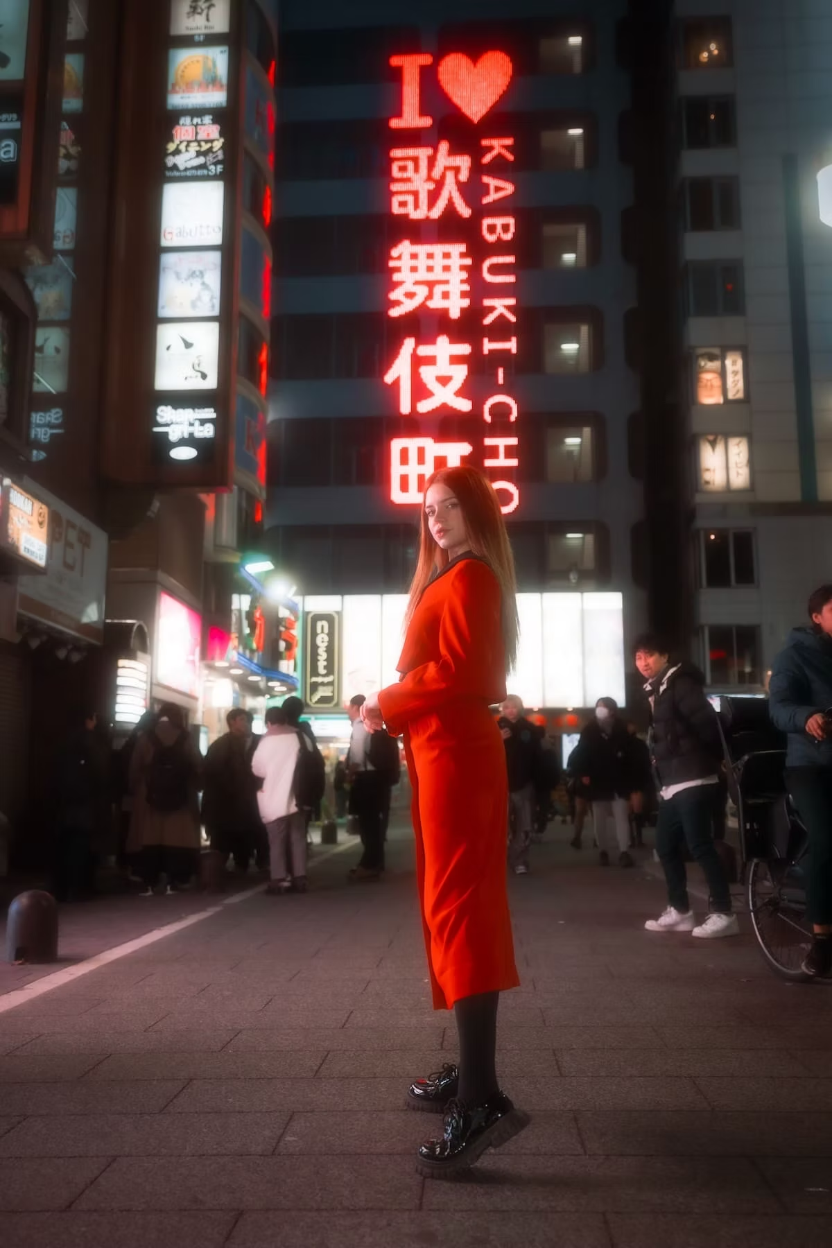 A woman in a red coat and black boots stands confidently in the vibrant Kabukicho area of Tokyo, surrounded by bright neon signs at night.