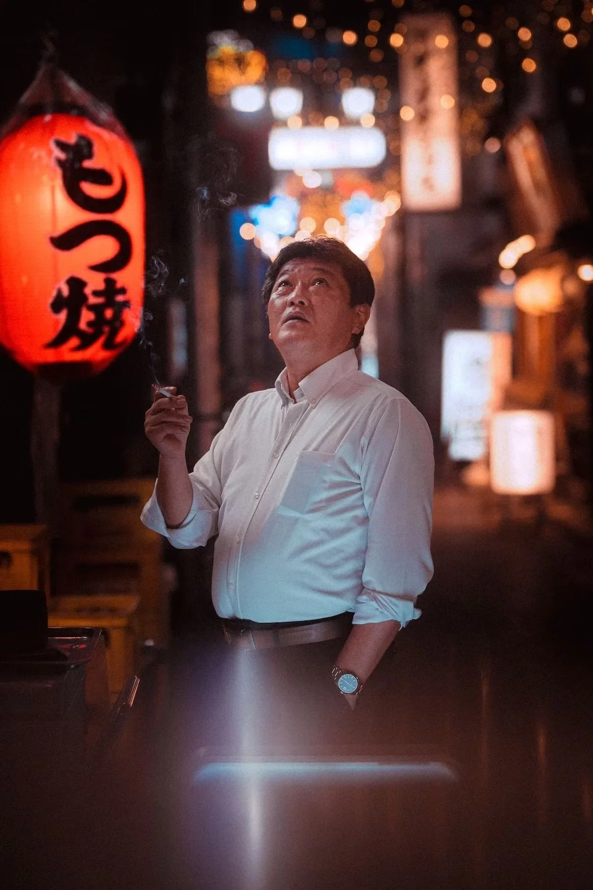 A Japanese man in a white shirt standing in front of a red paper lantern with black Japanese characters, in a dimly lit alley decorated with string lights at night.