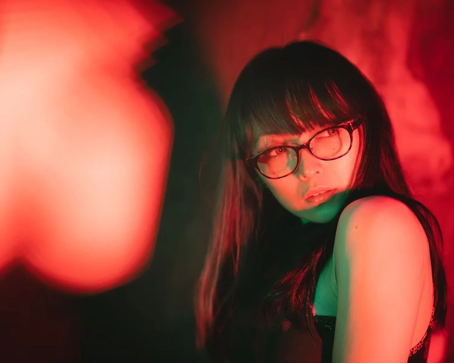 A woman with black hair and glasses posing in front of a red and green light background. Shot by Tokyo portrait photographer Deniz Demir.
