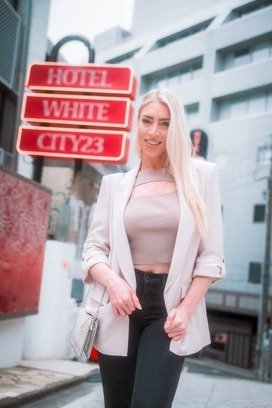A young woman with long blonde hair standing on a city street, smiling at the camera, wearing a light blazer and dark pants, with hotel signs in the background.