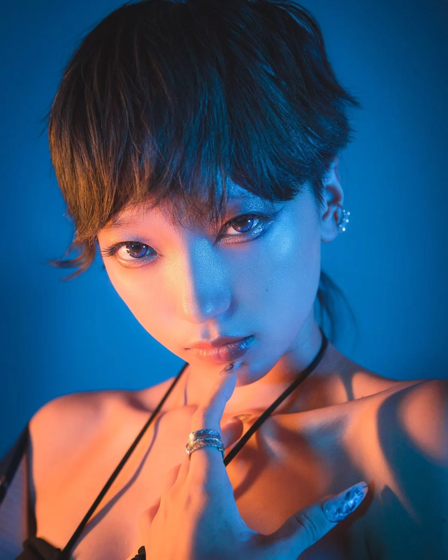 A close-up of a person with short dark hair, blue lighting on their face, and wearing silver jewelry, including rings and earrings, against a blue background.