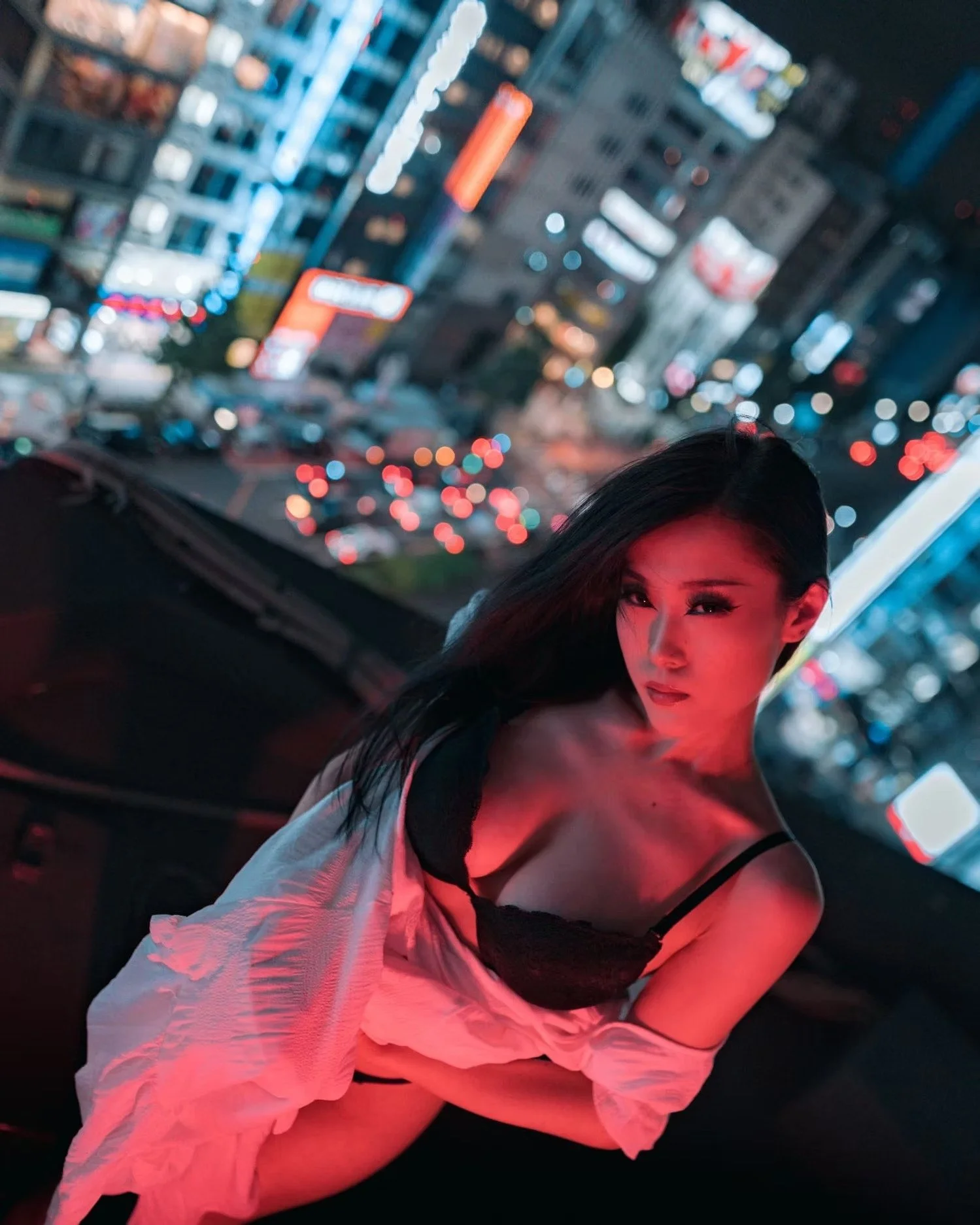 A woman with long dark hair and makeup, wearing a black and white outfit, sitting in a car at night with city lights blurred in the background on a Shinjuku Tokyo rooftop.