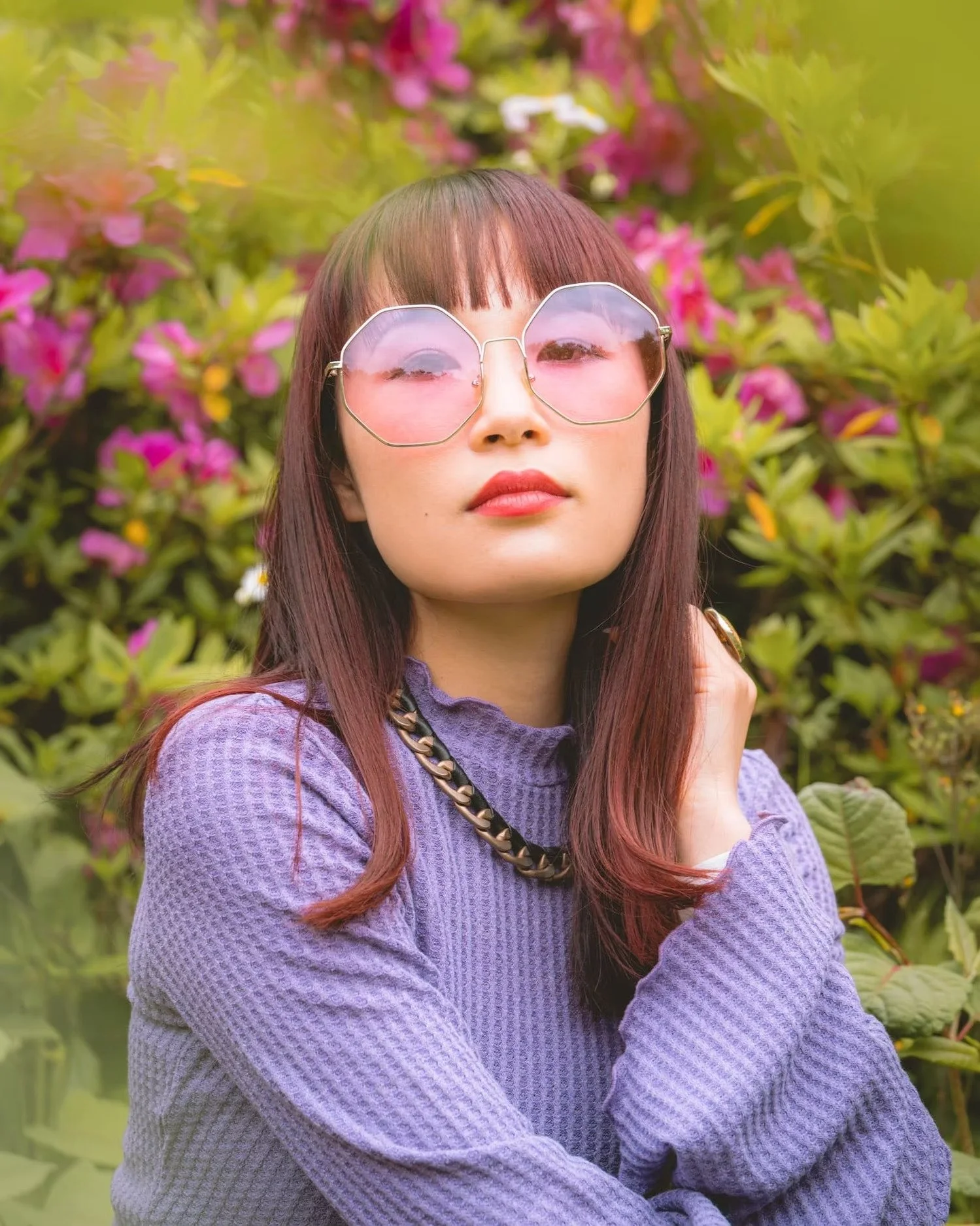 A woman with long, wavy brown hair with red highlights wearing large, heart-shaped pink sunglasses and a purple textured sweater, standing in front of pink and green foliage.