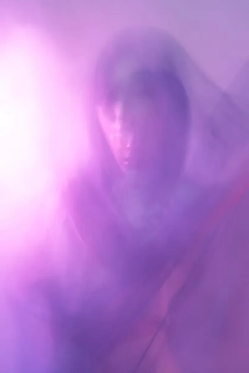 A person taking a mirror selfie with a purple-pink smudged filter, sunglasses, and dark hair in a dimly lit room.