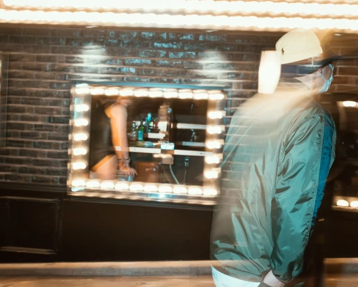 Person wearing a hat and face mask looking at a mirror framed with lights, with a brick wall in the background and bunny girls in the window in Shibuya.
