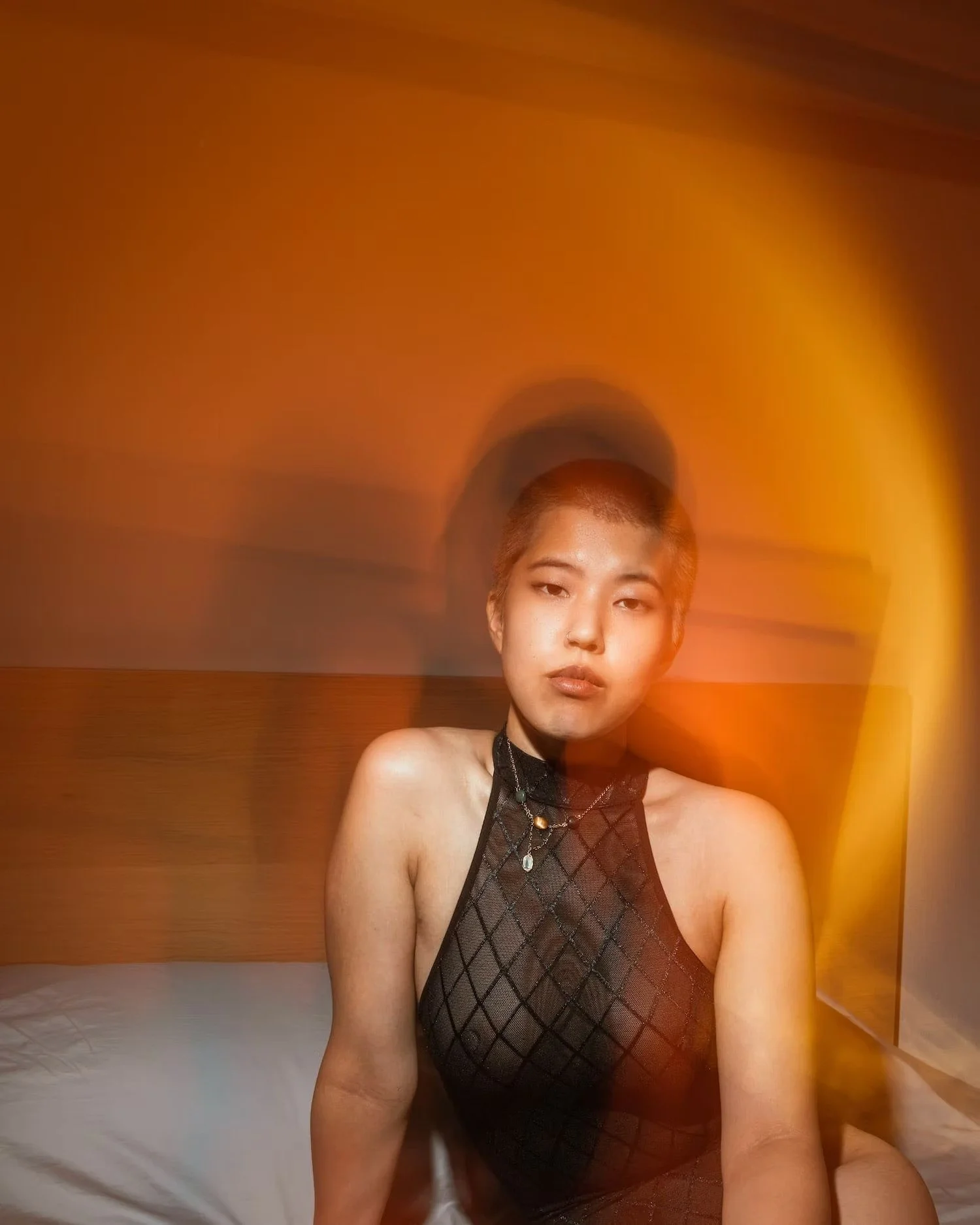 A young woman with short hair wearing a black lace halter top and layered necklaces sitting on a bed against an orange wall.