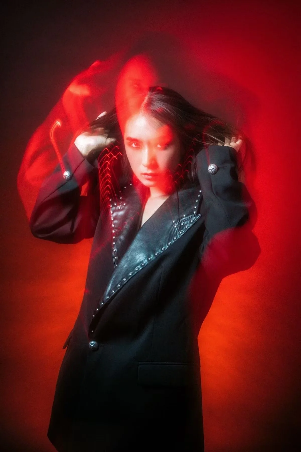 A woman with long dark hair wearing a black jacket with silver studs and buttons, posing against a red background with red light streaks.