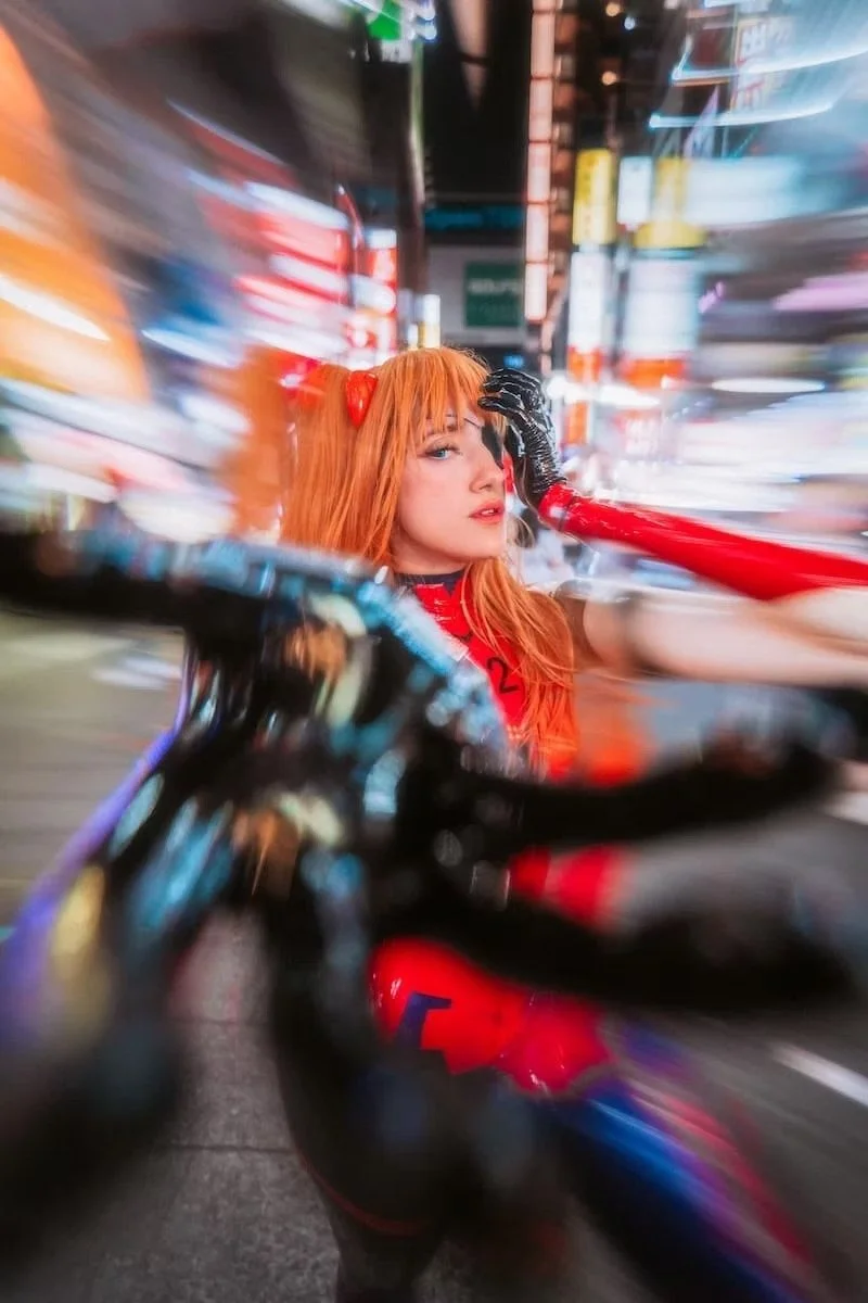 A woman with red hair and a red bow, dressed in a red and black costume, standing on a city street at night with motion blur effects surrounding her. Asuka cosplay from Evangelion.