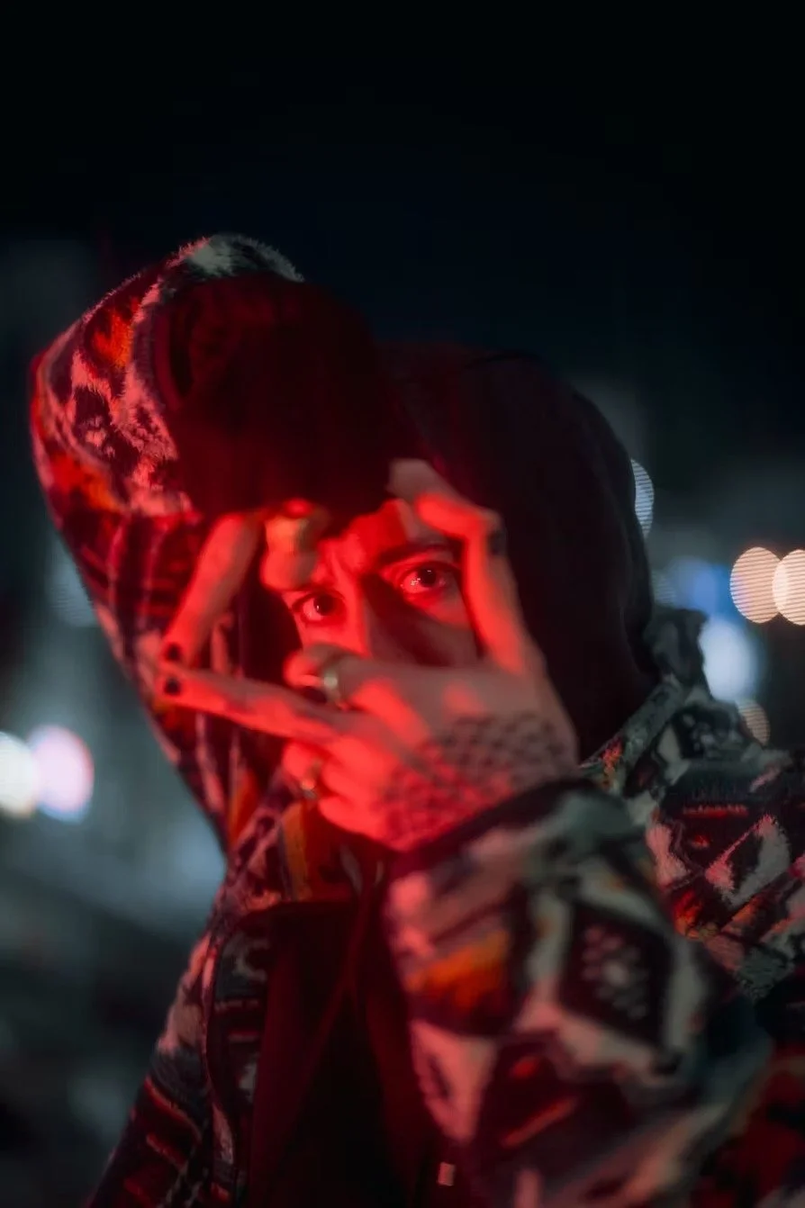A young person with dark hair in a hooded jacket making a hand gesture near their face, illuminated by red and ambient city lights at night.