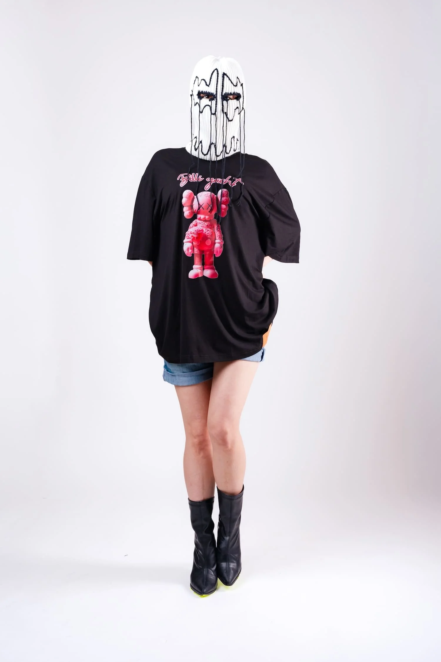 Person wearing a black oversized t-shirt with a pink graphic design, denim shorts, black boots, and a white mask with black line art design hiding their face, standing against a plain white background.