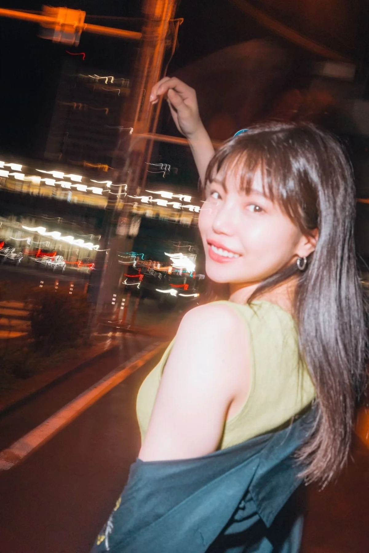 A young woman smiles as she looks back over her shoulder, standing under the bright lights of a Tokyo street at night. The motion blur of the background enhances the sense of movement and excitement. Street photography by Deniz Demir.