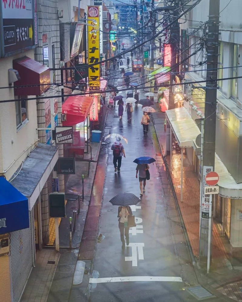 People walking with umbrellas on a rainy street lined with storefronts, signs, and wires overhead.