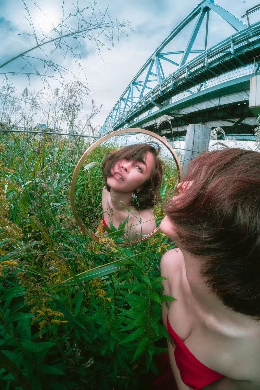 A woman with short brown hair looking into a round mirror amidst tall green grass and plants outdoors, with a large blue bridge in the background. Fish eye photography by Deniz Demir.