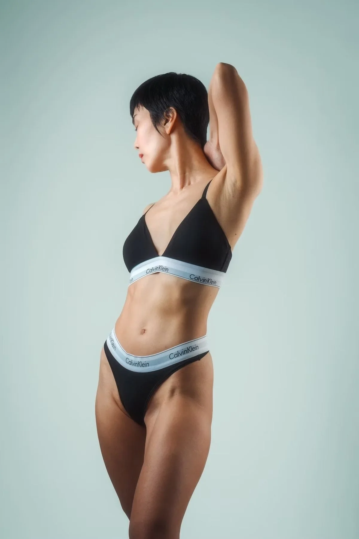 A woman with short black hair wearing black Calvin Klein underwear, standing sideways with one arm raised behind her head against a plain light background.