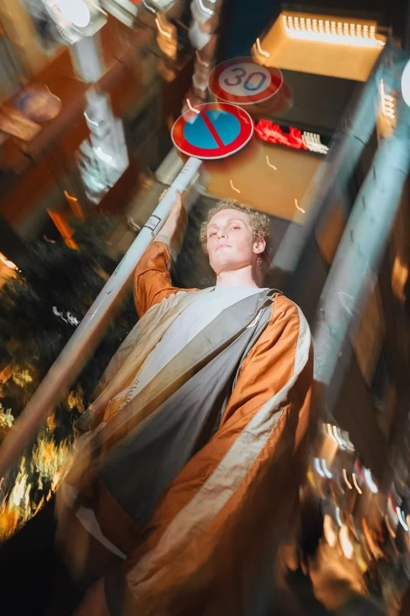 A young man stands in a busy street at night surrounded by blurred lights, signs, and buildings. He looks directly at the camera with a confident expression, holding a pole with a no-parking sign and a speed limit sign above his head.
