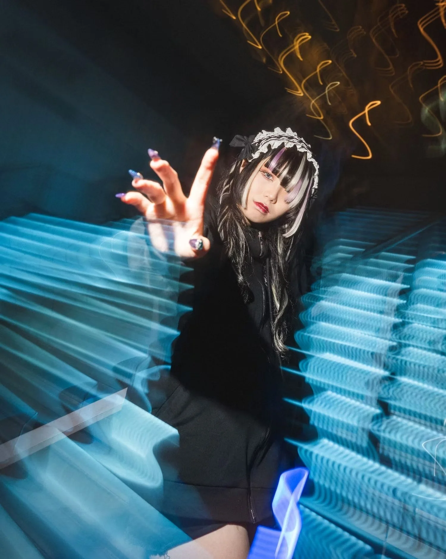 A young Japanese woman with black and white hair, wearing a black hoodie with the hood up, is making a peace sign with her right hand. She has colorful nails and is standing in front of a background with blue and yellow streaks of light, creating a m