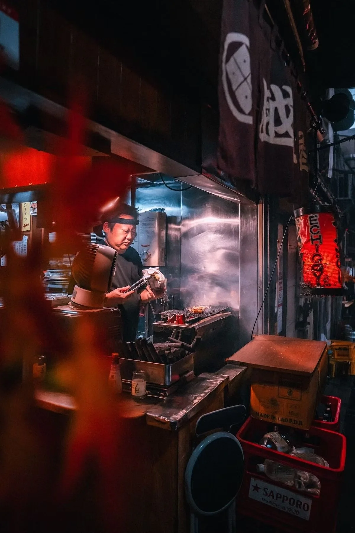 A man preparing food in a small, dimly lit Japanese street food stall at night. The man is wearing a black shirt and headlamp, cooking over a grill with smoke rising. The stall has Japanese signage, with various cooking utensils and supplies visible.