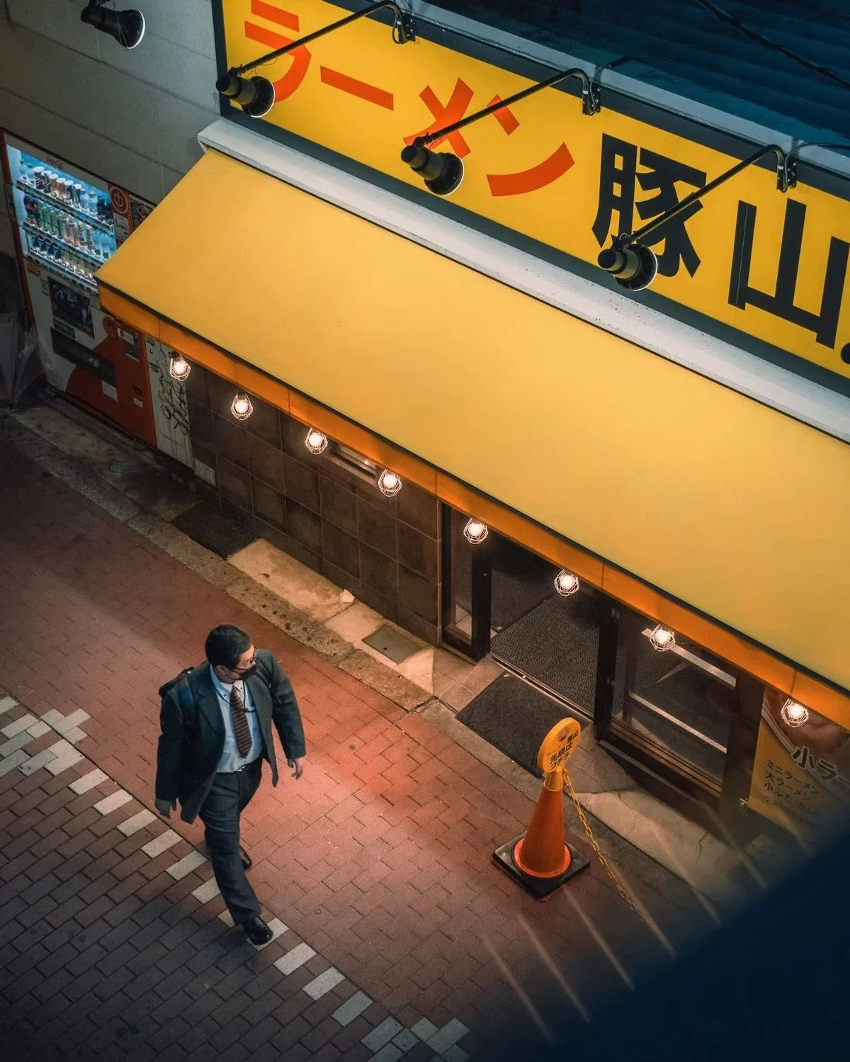 A man wearing a suit and face mask walking on a brick sidewalk outside a ramen store with a yellow awning and orange traffic cone.