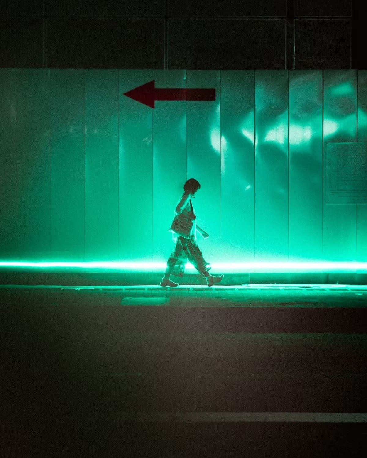 Person walking on a sidewalk in front of a wall illuminated with green neon lights, with a red arrow sign pointing left in Nagasaki.