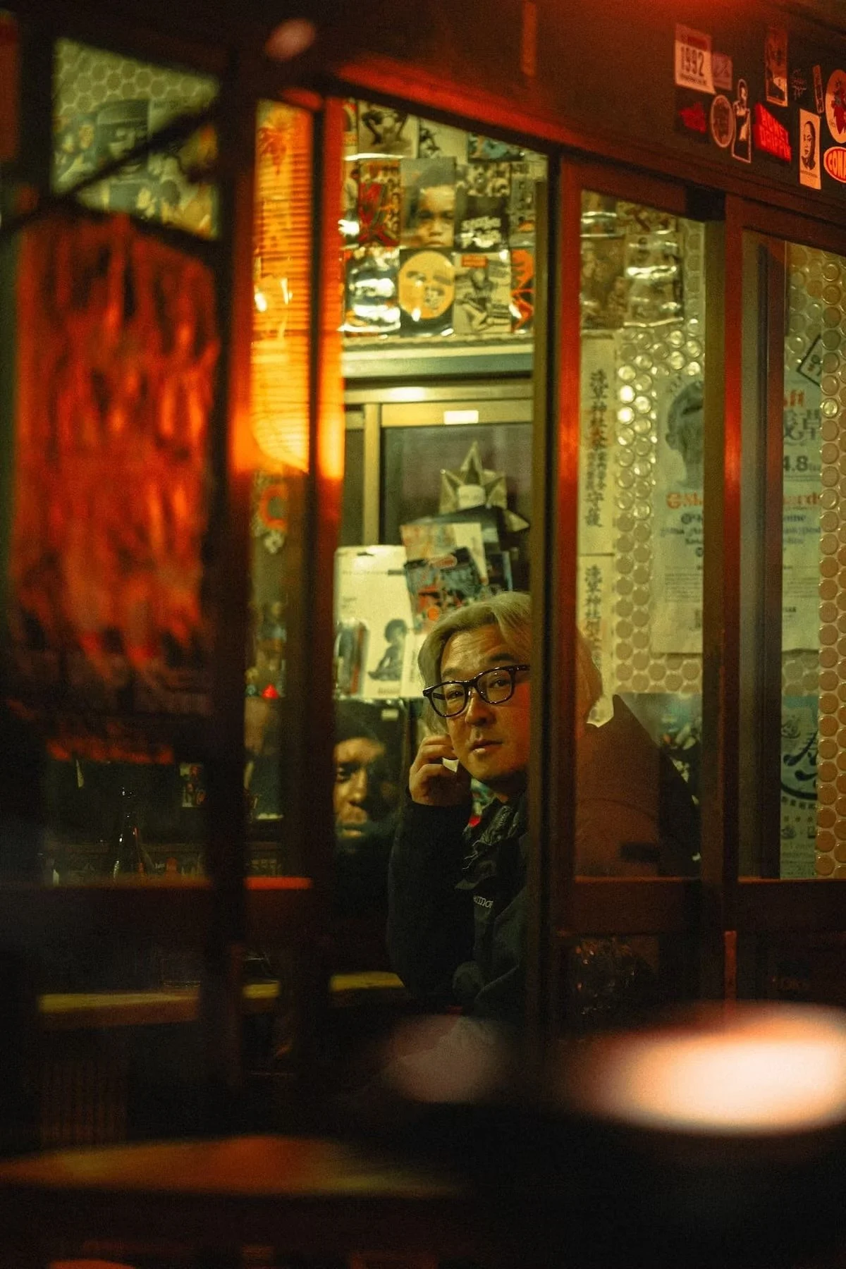 A Japanese person with gray hair and glasses looking through a window at another person seated indoors, surrounded by posters and decorations in a dimly lit setting in Ueno.