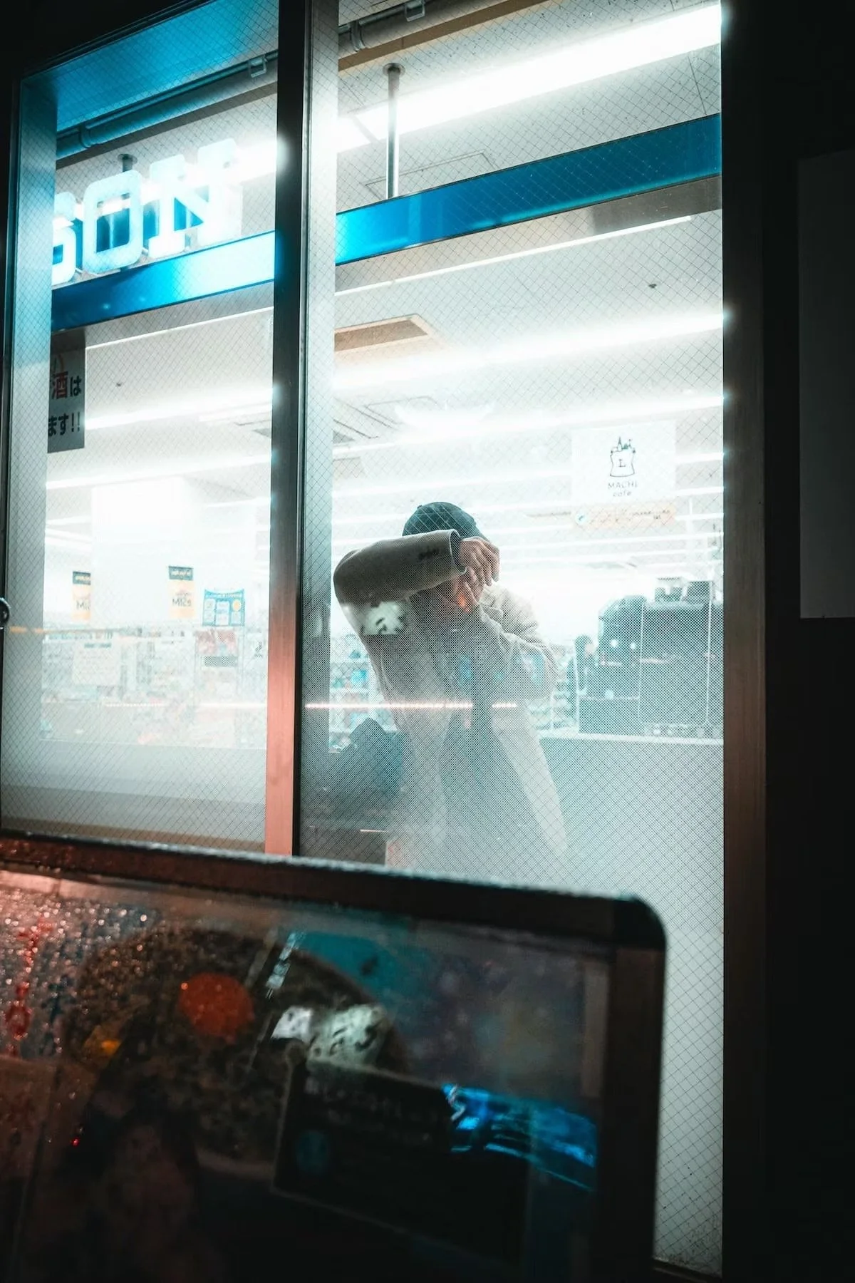 Reflection of a person taking a selfie in a store window. The store, possibly a convenience store, has bright fluorescent lighting and visible shelves with products inside. The photo was taken from inside a vehicle, as part of a dashboard is visible 