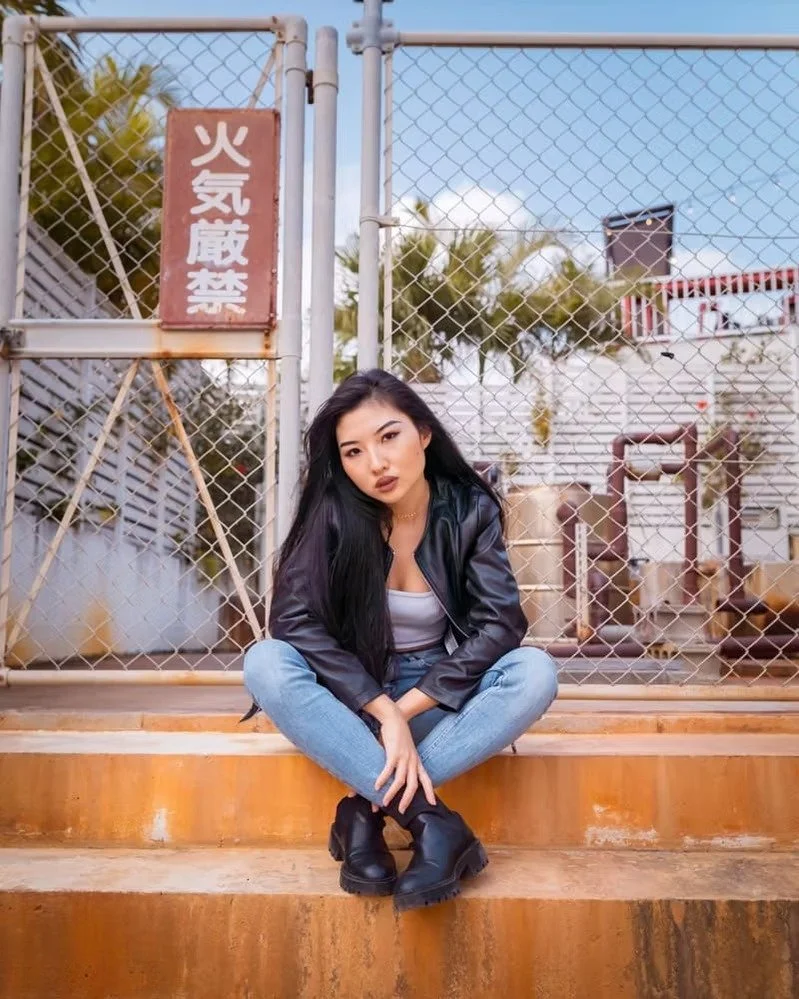 A young woman with long black hair, wearing a black leather jacket, gray tank top, blue jeans, and black boots, sitting cross-legged on concrete steps in front of a chain-link fence with a red Japanese warning sign, under a partly cloudy sky.