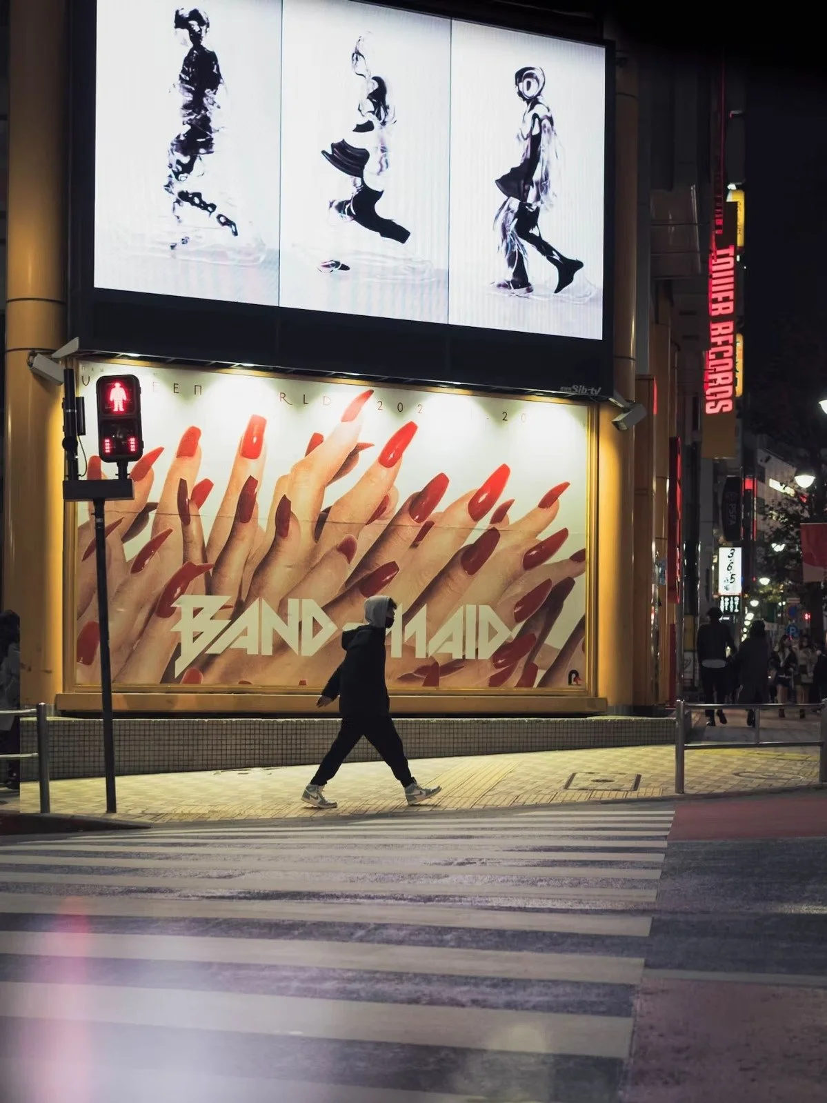 Shibuya street scene at night with large illuminated billboards, including one advertising a band called 'Band-Maid' with a close-up of manicured red nails. A person wearing a black hoodie walks across a crosswalk, and there is a pedestrian signal wi