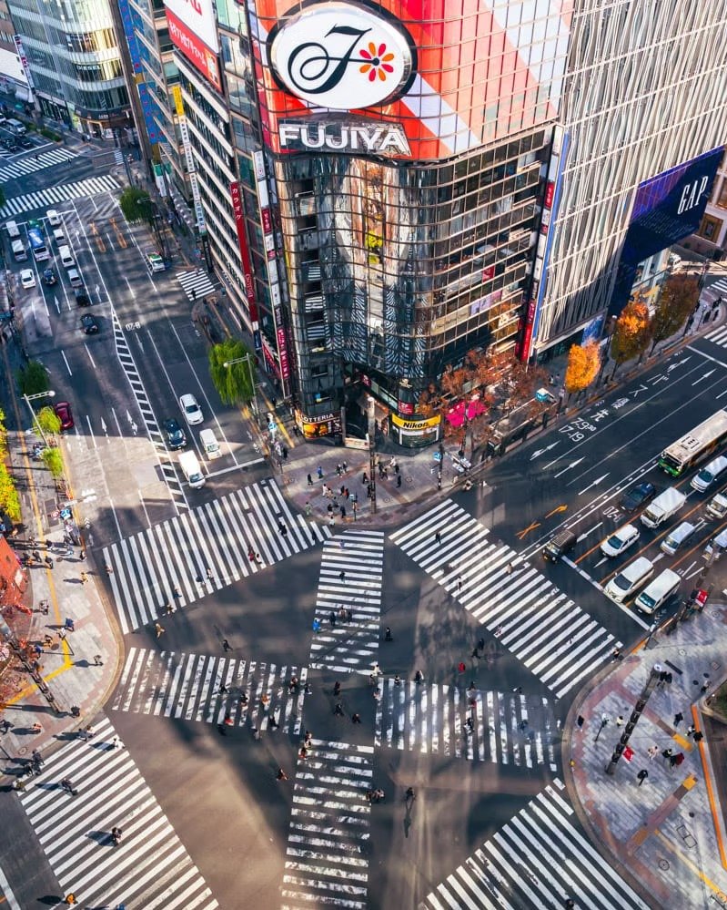 Aerial view of a busy city intersection with crosswalks and pedestrians, surrounded by tall buildings with illuminated signs.