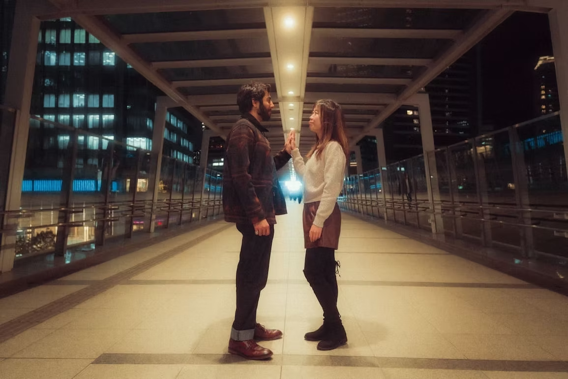 A couple faces each other in a well-lit outdoor walkway in Tokyo. They are standing in front of large glass windows, with their eyes locked and a playful interaction.