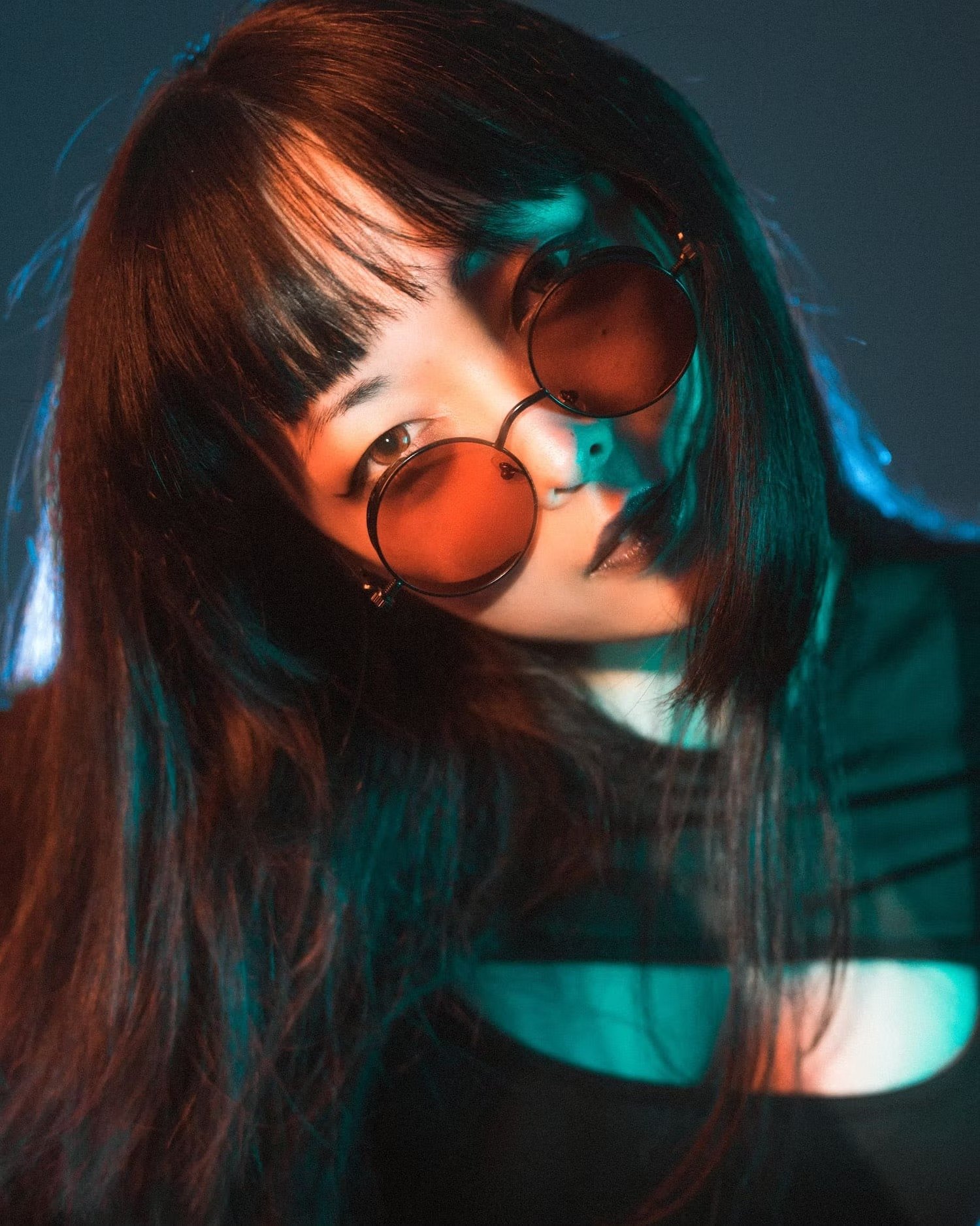 A woman with dark hair and bangs wearing round sunglasses with tinted lenses, looking at the camera with a subtle expression. The lighting has a mix of warm and cool tones, creating a dramatic effect.