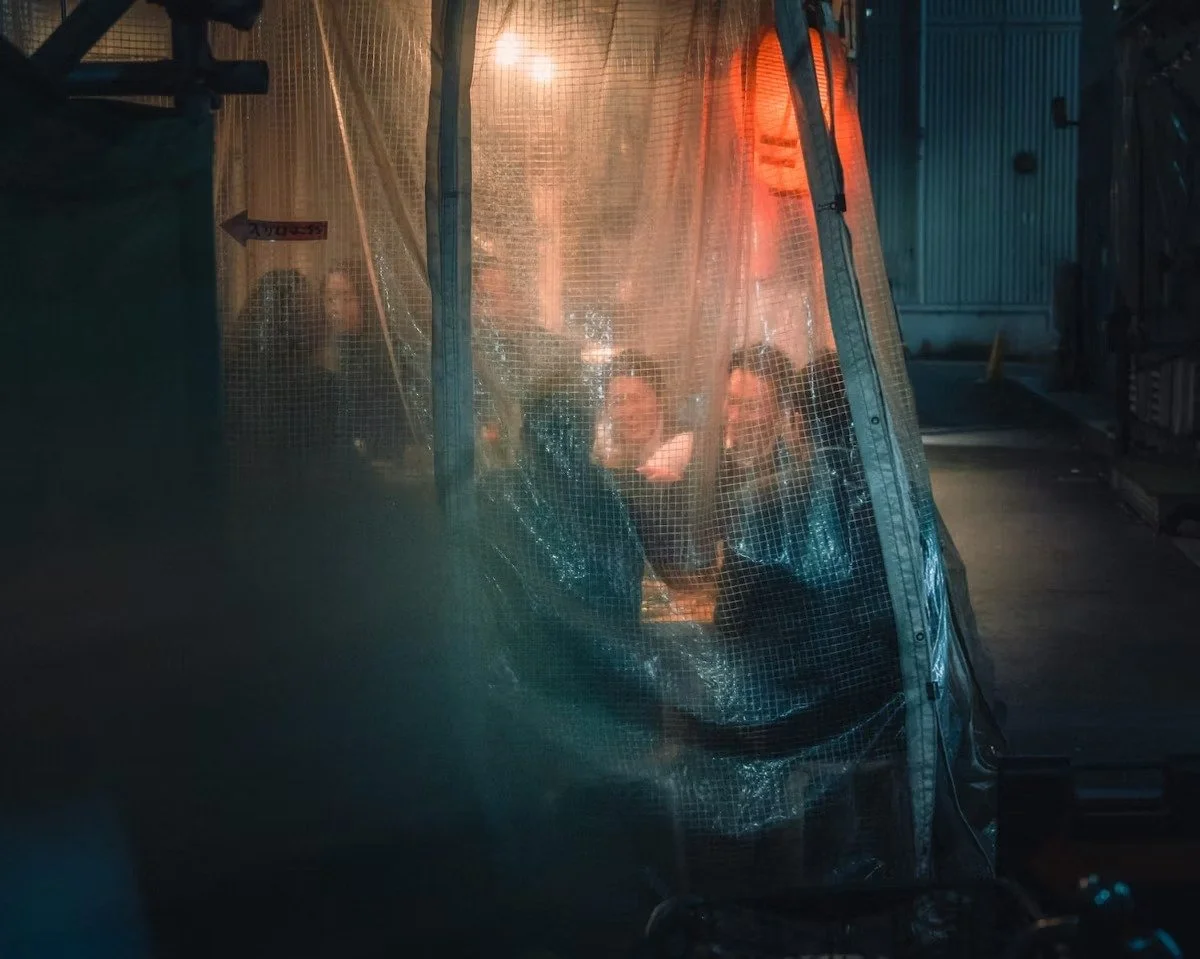 People sitting and smiling inside a transparent tent or booth, illuminated by a warm light, in an outdoor setting at night in an Izakaya.