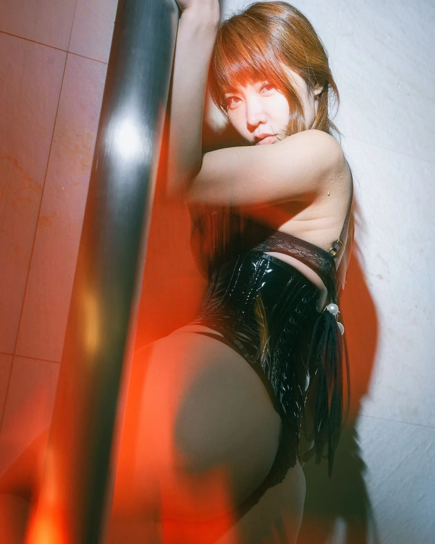 A woman with auburn hair, wearing a black shiny dress, is leaning against a wall with her eyes looking into the camera. The photo is taken from a low angle with warm lighting and a partially visible metal pipe in the foreground.