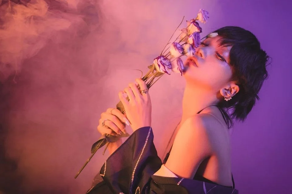 A woman in a black bikini stands with a bouquet of flowers in her hand, surrounded by colorful smoke. The lighting casts a warm, moody glow, highlighting her confident expression and adding to the artistic, bold feel of the image.