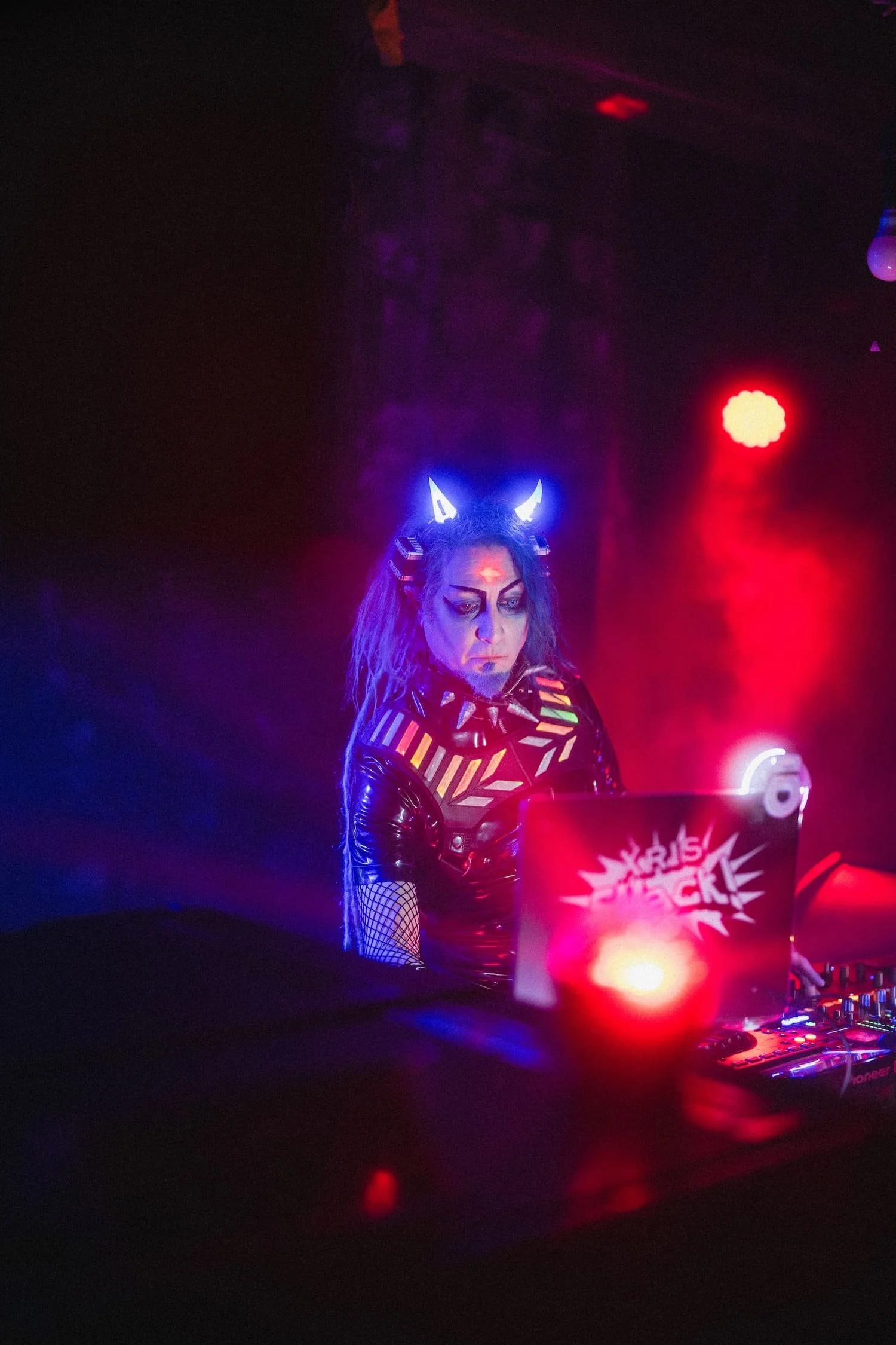 Bondage Ball 
---
A woman dressed as a neon-lit demon DJ with glowing horns, wearing a colorful geometric top and fishnet sleeves, working at a DJ booth in a dark, club-like setting with red and purple lighting.