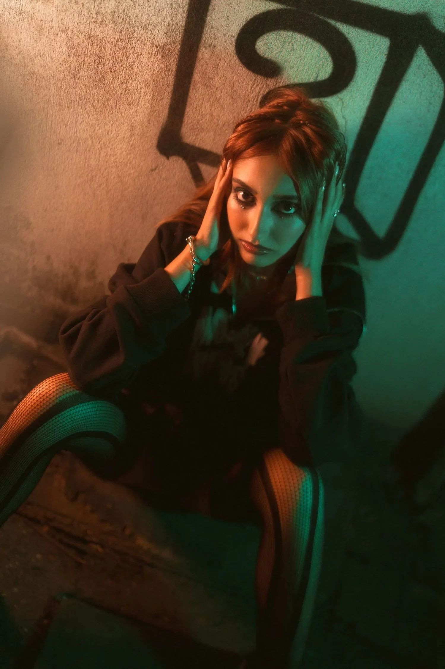 A woman with red hair sat on the ground, resting her head in her hands near a wall with a large black-edged geometric design. She wears black clothing, fishnet stockings with colorful rainbow stripes, and silver jewelry. The lighting creates a moody 