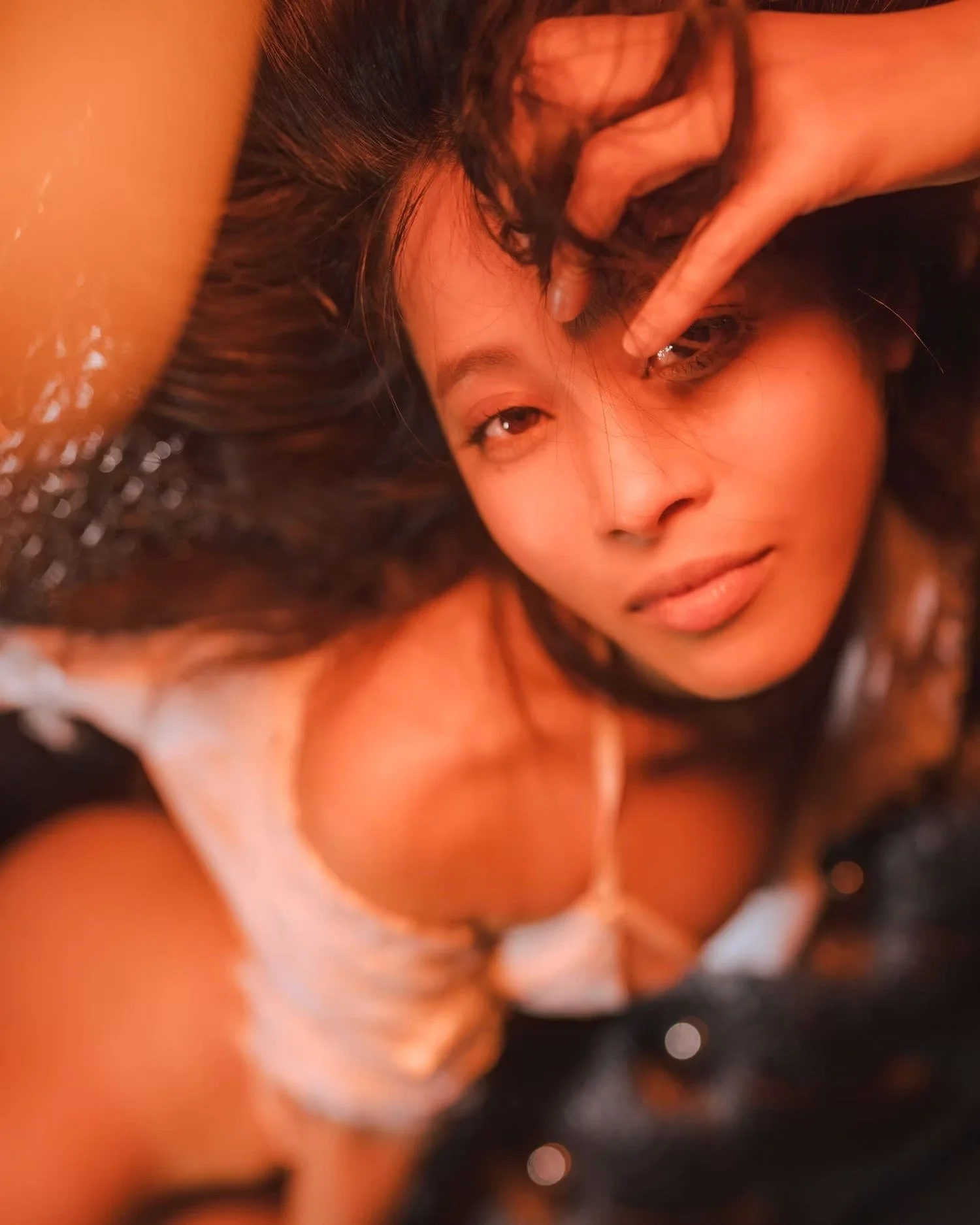 A close-up of a woman with dark curly hair and light skin, making a playful face with her lips slightly parted and eyes looking into the camera. She is wearing a white top with thin straps.