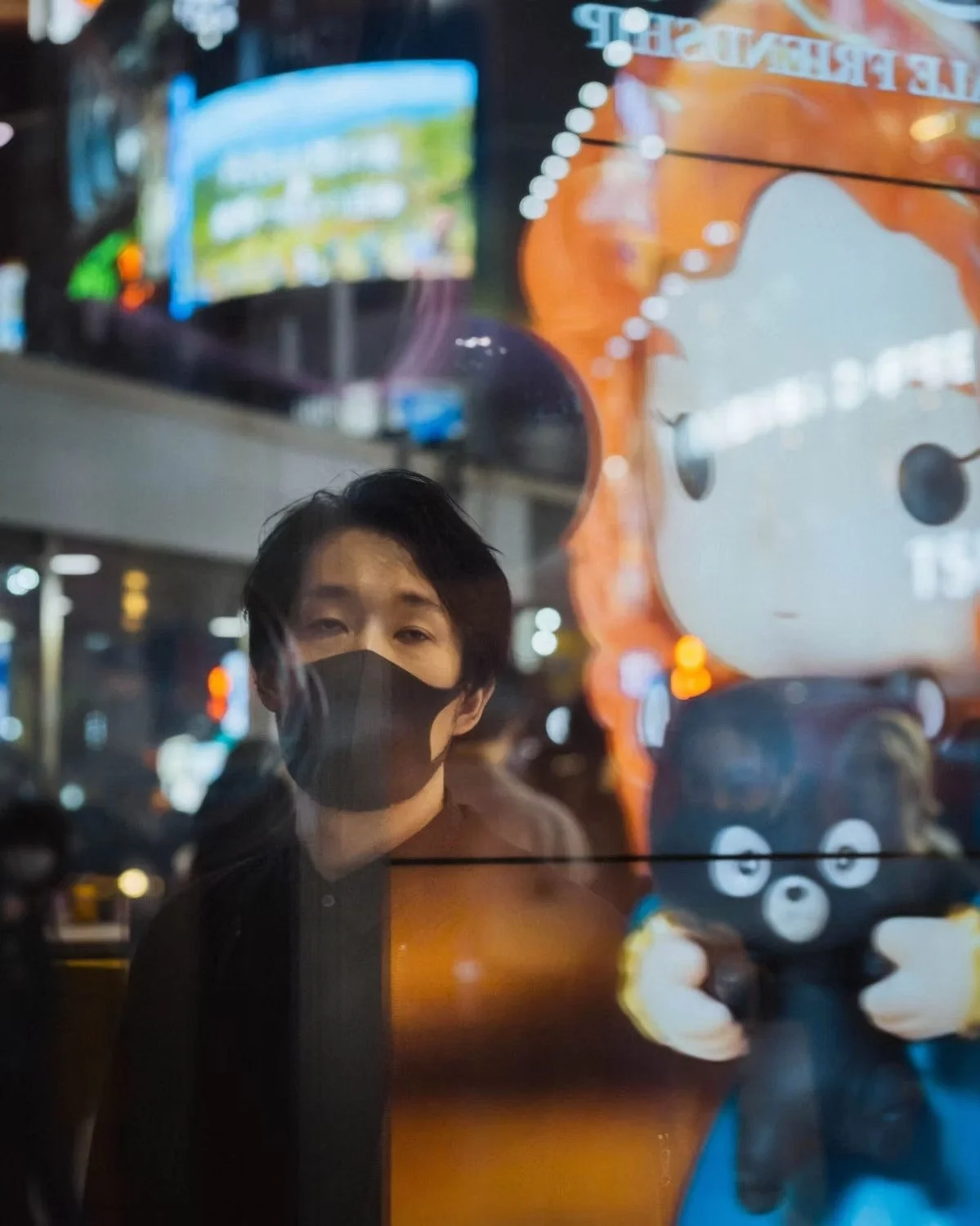 Reflection of a young man with black hair and a black face mask on a glass window, with a cartoon character plush toy and city lights in the background in Shibuya.