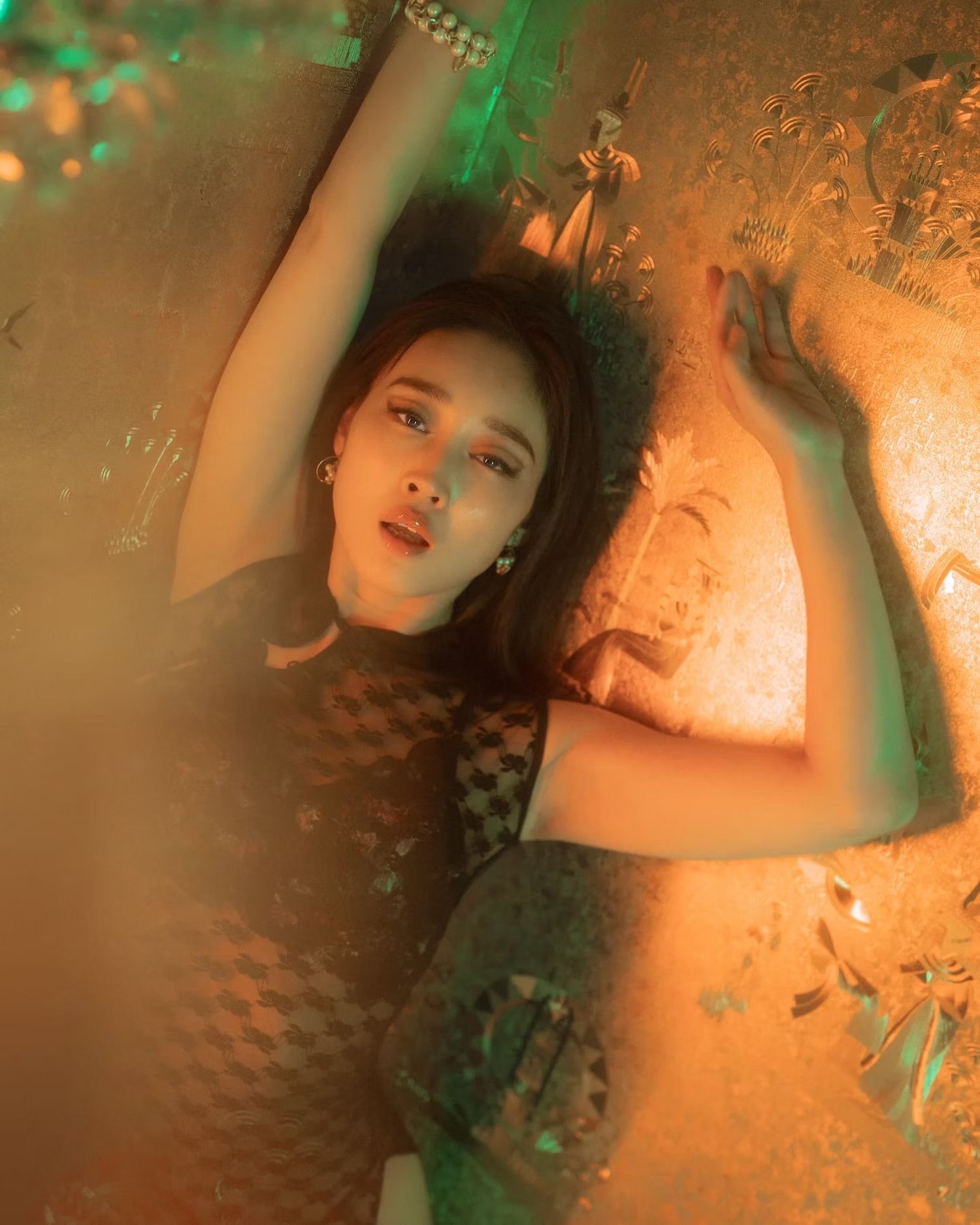 A woman with long brown hair lying on the floor with colorful lights and confetti around her, wearing a black dress with a dark floral pattern and earrings.