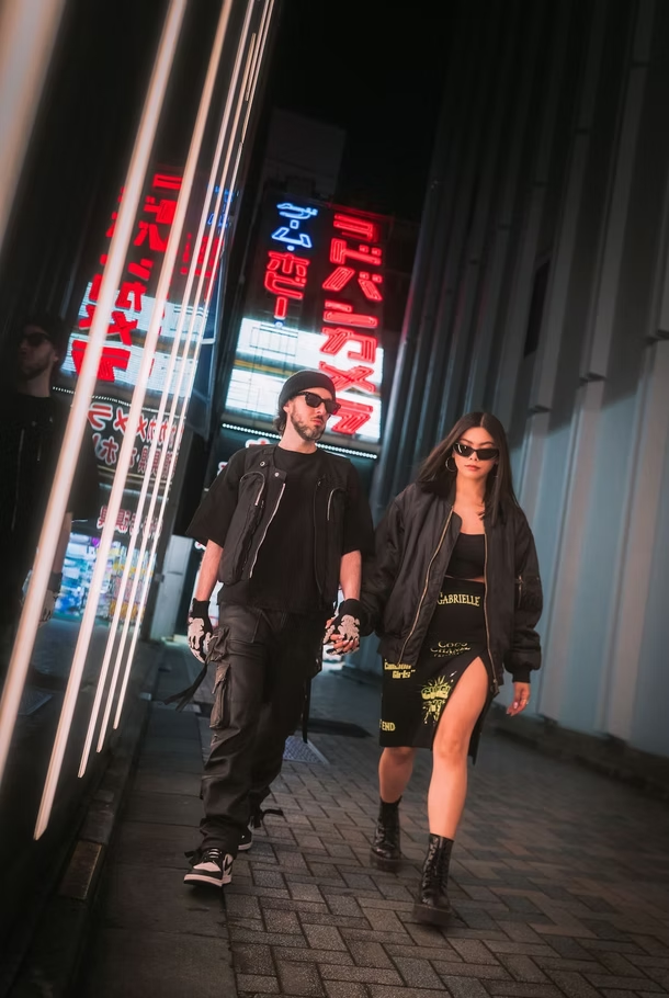 Two stylish young adults with dark hair and sunglasses walking on a city sidewalk at night, dressed in black with neon signs in Japanese characters reflecting on glass windows.