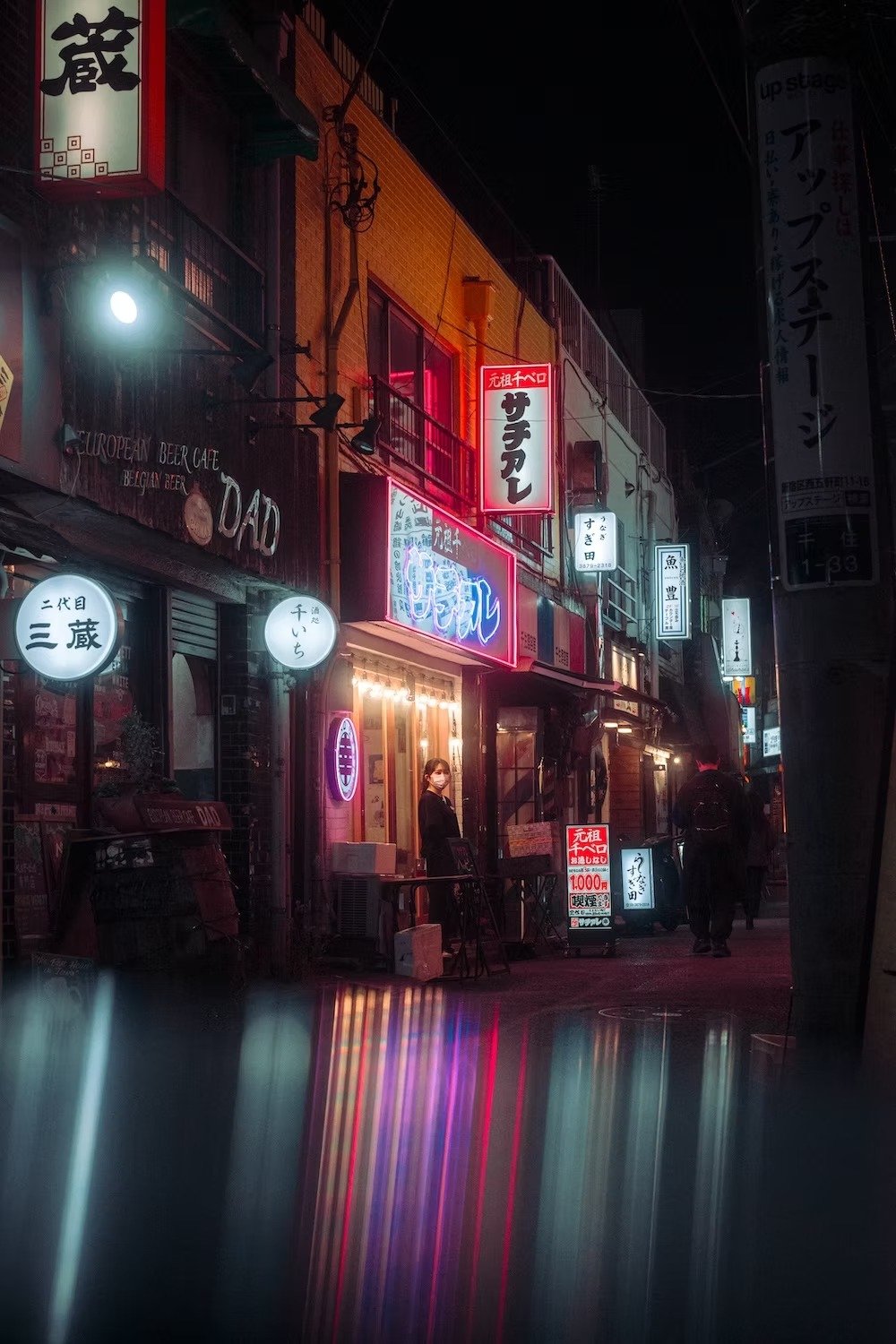 Nighttime street scene with illuminated Japanese signs and a person standing outside a restaurant.