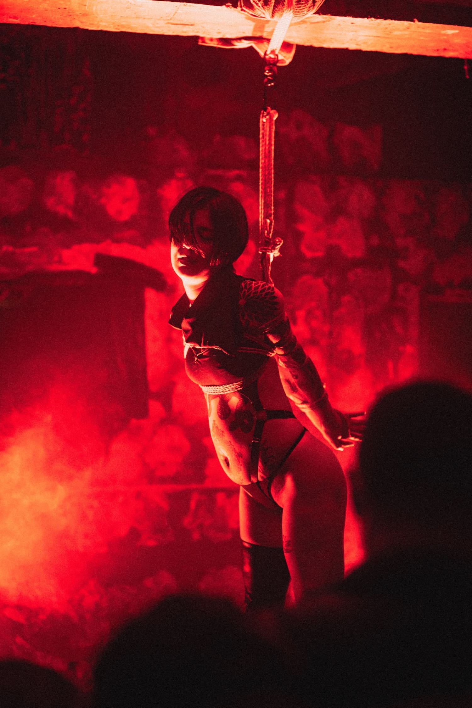 Bondage Ball 
---
A woman is kneeling and tied with a rope in a dimly lit room with red lighting. The background features a red patterned wall.
