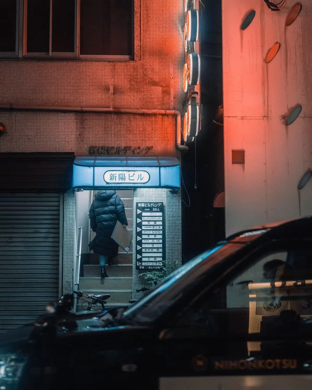 Person entering a building with a blue illuminated sign, Japanese text on the sign, and a signage board outside with multiple listings, night scene with vehicles parked in front, and orange and pink lighting Shimbashi nights.